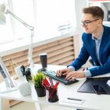 Junger Mann mit Brille arbeitet an einem modernen Schreibtisch. Er ist mit Hemd und Blazer bekleidet, vor ihm auf dem Tisch stehen ein Monitor, kleine Lautsprecherboxen, ein Wecker, eine Pflanze und ein Stifthalter. Handy, Kalender und ein Kaffeebecher. Die Einrichtung des Büros ist hell und freundlich. 