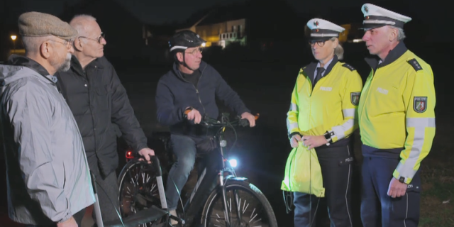 Zwei Beamte stehen neben einem Fahrradfahrer. Neben ihm stehen zwei weitere Herren. Es ist dunkel.
